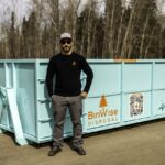 Man stands beside a large turquoise dumpster labeled "BinWise Disposal" with a QR code. He's wearing sunglasses and a black shirt with an orange