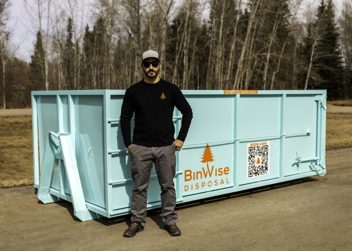 Man stands beside a large turquoise dumpster labeled "BinWise Disposal" with a QR code. He's wearing sunglasses and a black shirt with an orange