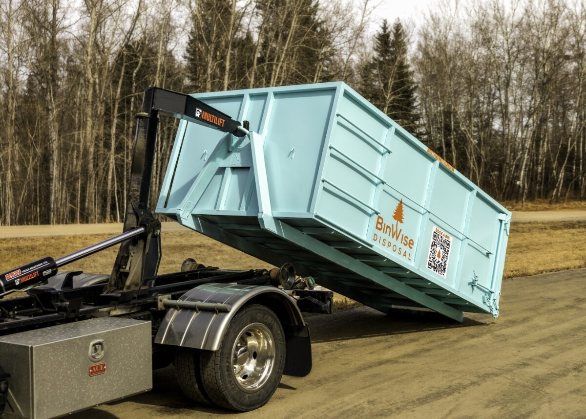 A turquoise disposal bin being unloaded by a truck with a Multilift mechanism on a rural dirt road, surrounded by bare trees.