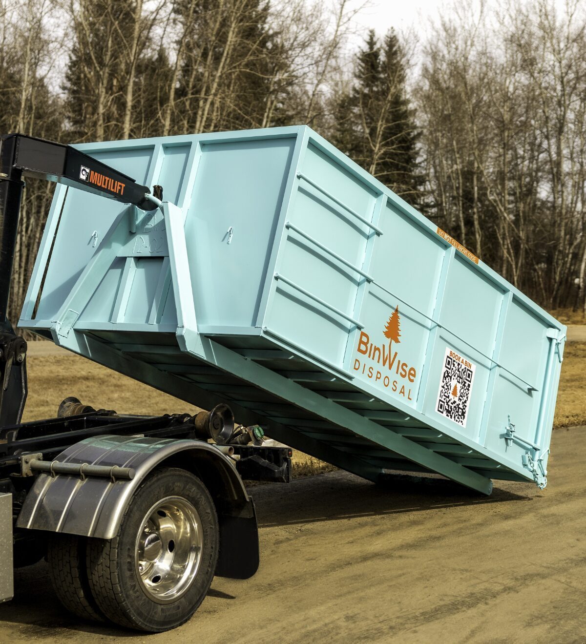 A turquoise dumpster is being unloaded from a truck with the logo "BinWise Disposal." It's set against a backdrop of bare trees on a clear day.