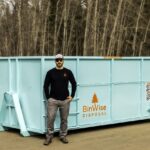 A man in sunglasses and a cap stands beside a large teal disposal bin labeled "BinWise Disposal" with a QR code, set outdoors near a wooded area.