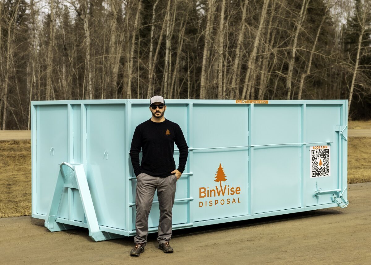 A man in sunglasses and a cap stands beside a large teal disposal bin labeled "BinWise Disposal" with a QR code, set outdoors near a wooded area.