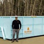 A man in sunglasses and a hat stands in front of a large aqua-colored disposal bin labeled "BinWise Disposal" with a tree logo and QR code.