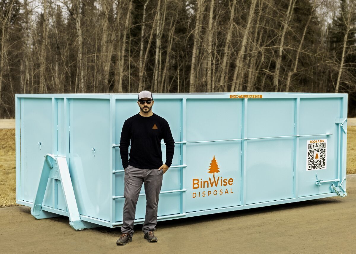 A man in sunglasses and a hat stands in front of a large aqua-colored disposal bin labeled "BinWise Disposal" with a tree logo and QR code.