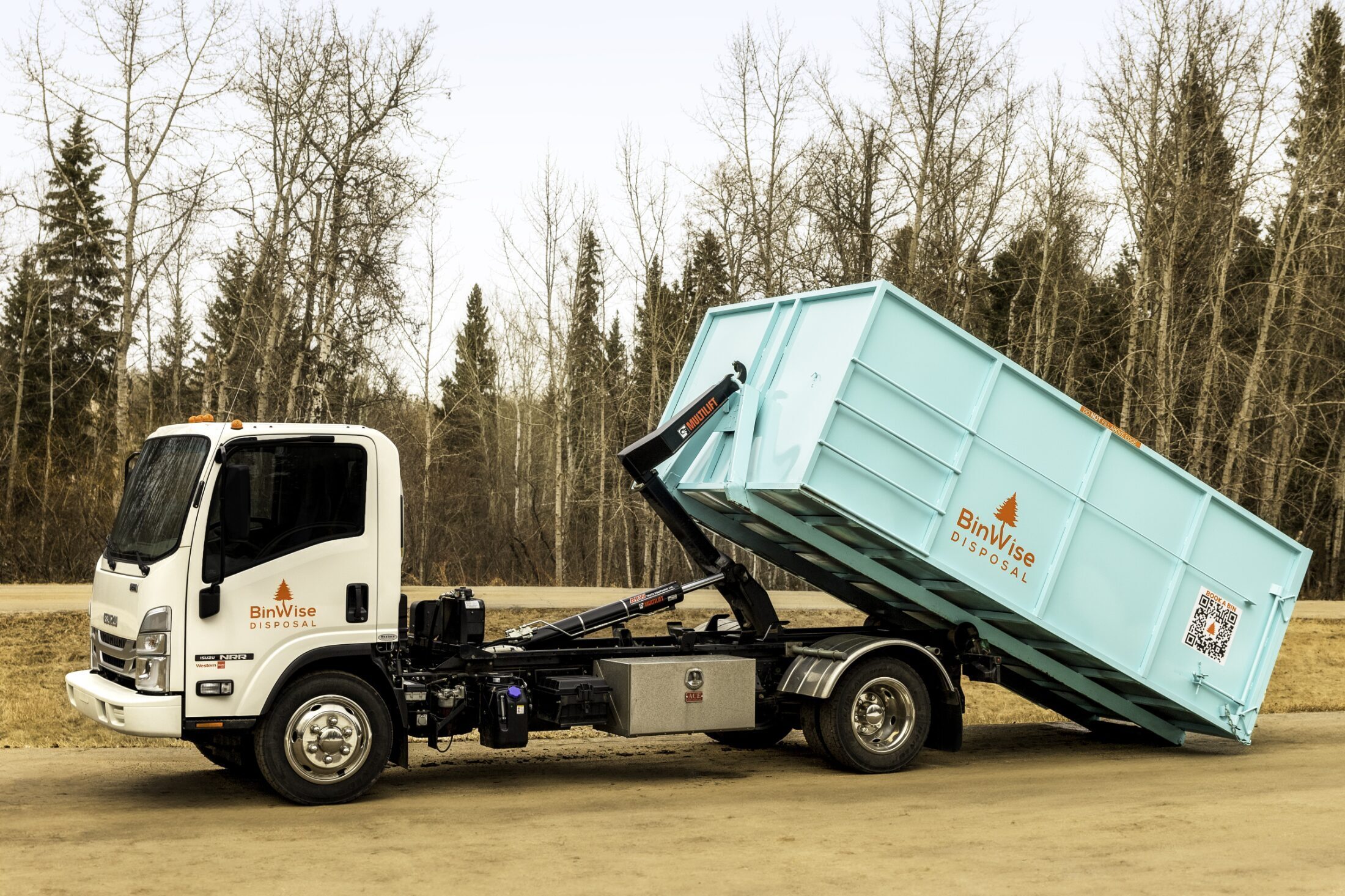 A white truck with "BinWise Disposal" logo is unloading a large teal container using a hydraulic arm. Trees and a dirt path are in the background.