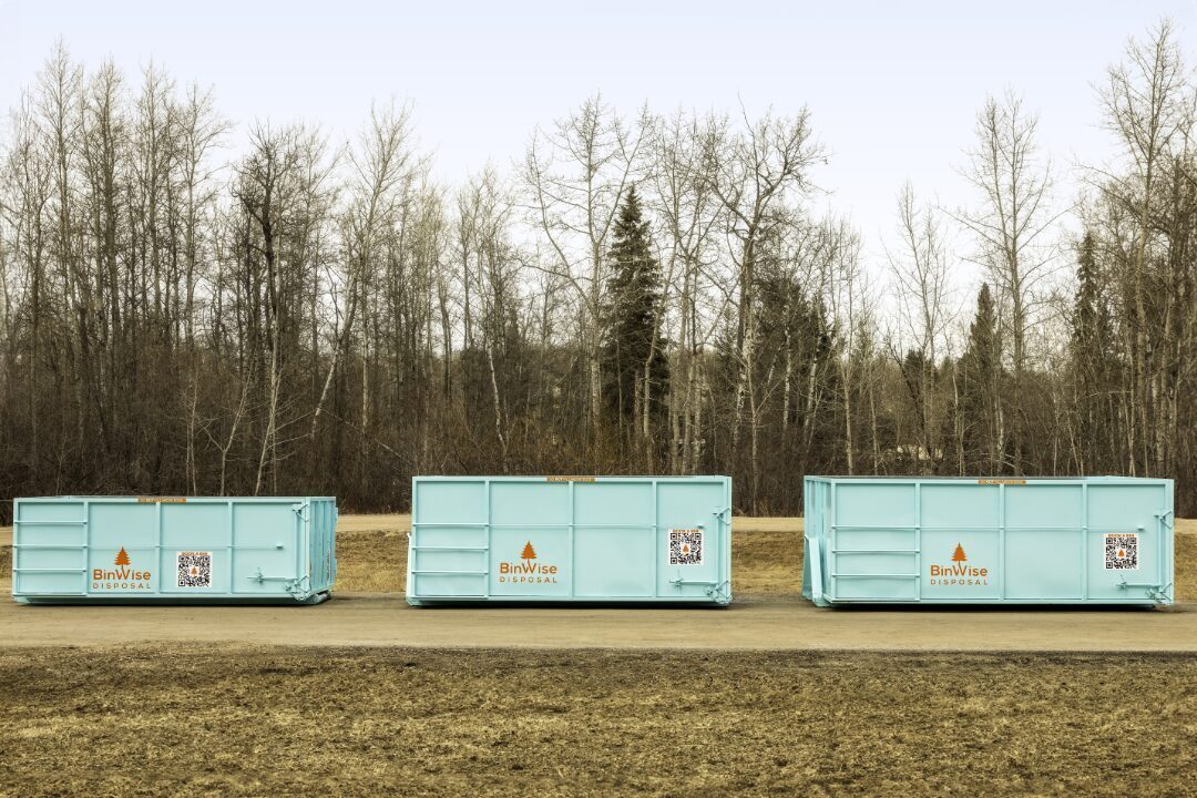 Three turquoise waste disposal bins labeled "BinWise Disposal" are lined up on a dirt road, with bare trees in the background.