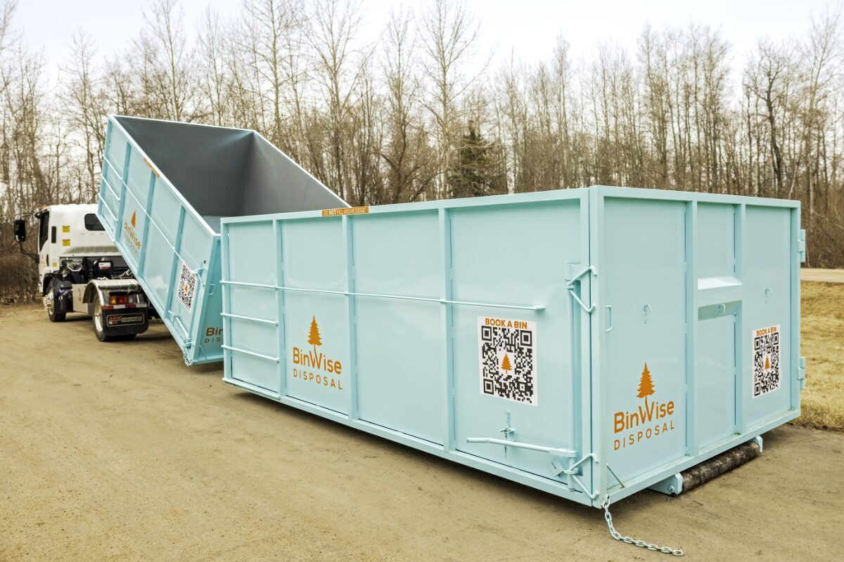 A turquoise dumpster with "BinWise Disposal" branding and QR codes is being unloaded from a white truck on a dirt road, surrounded by bare trees.
