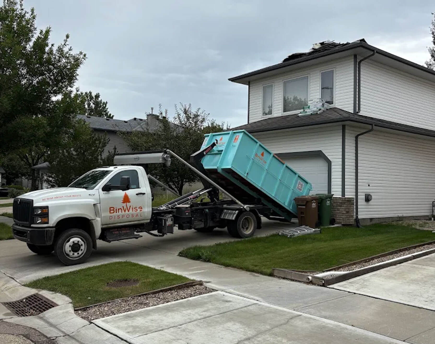 A turquoise dumpster with "BinWise Disposal" branding and QR codes is being unloaded from a white truck on a dirt road, surrounded by bare trees.
